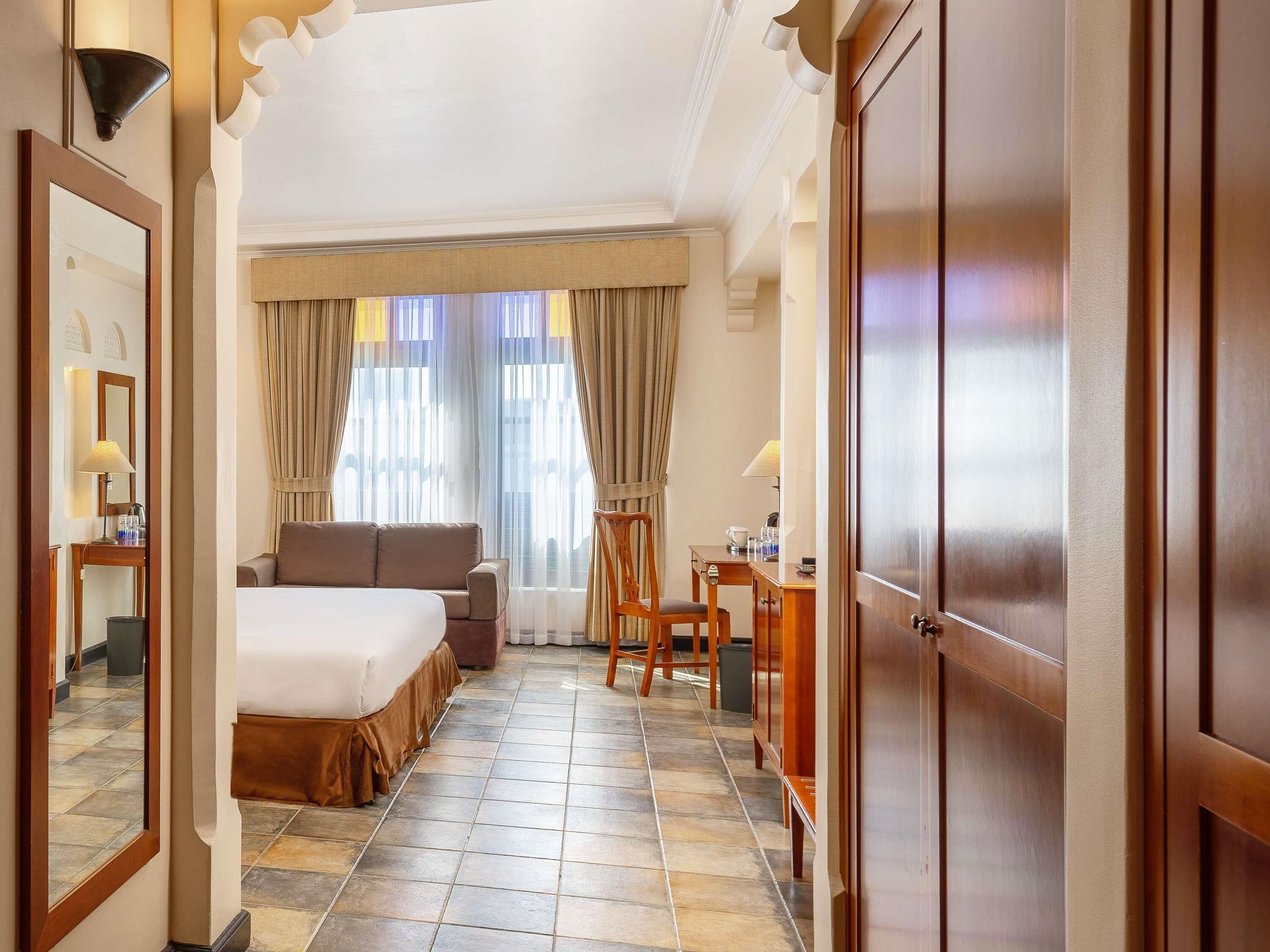 Ein Hotelzimmer mit Bett, Schreibtisch, Stuhl, Spiegel und einem Fenster mit Vorhängen.
