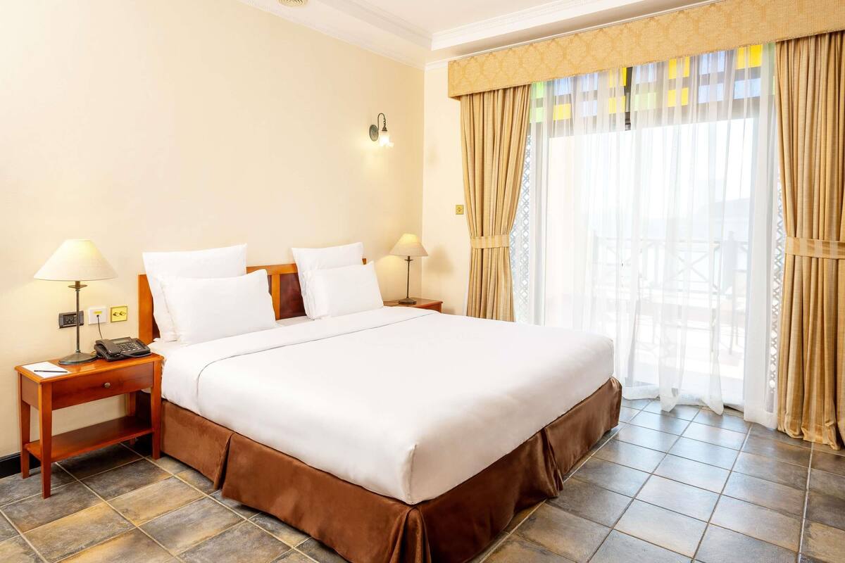 A hotel room with a large bed, two bedside tables with lamps, a telephone, and a view of the outdoors through sheer curtains.