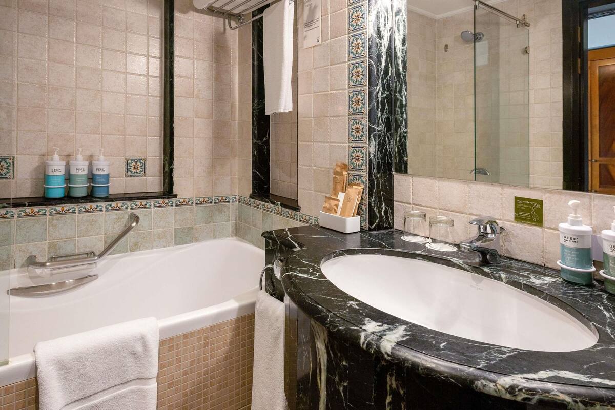 A bathroom with a bathtub, a sink with a marble countertop, and a mirror. There are toiletries on the countertop and a towel hanging on the wall.