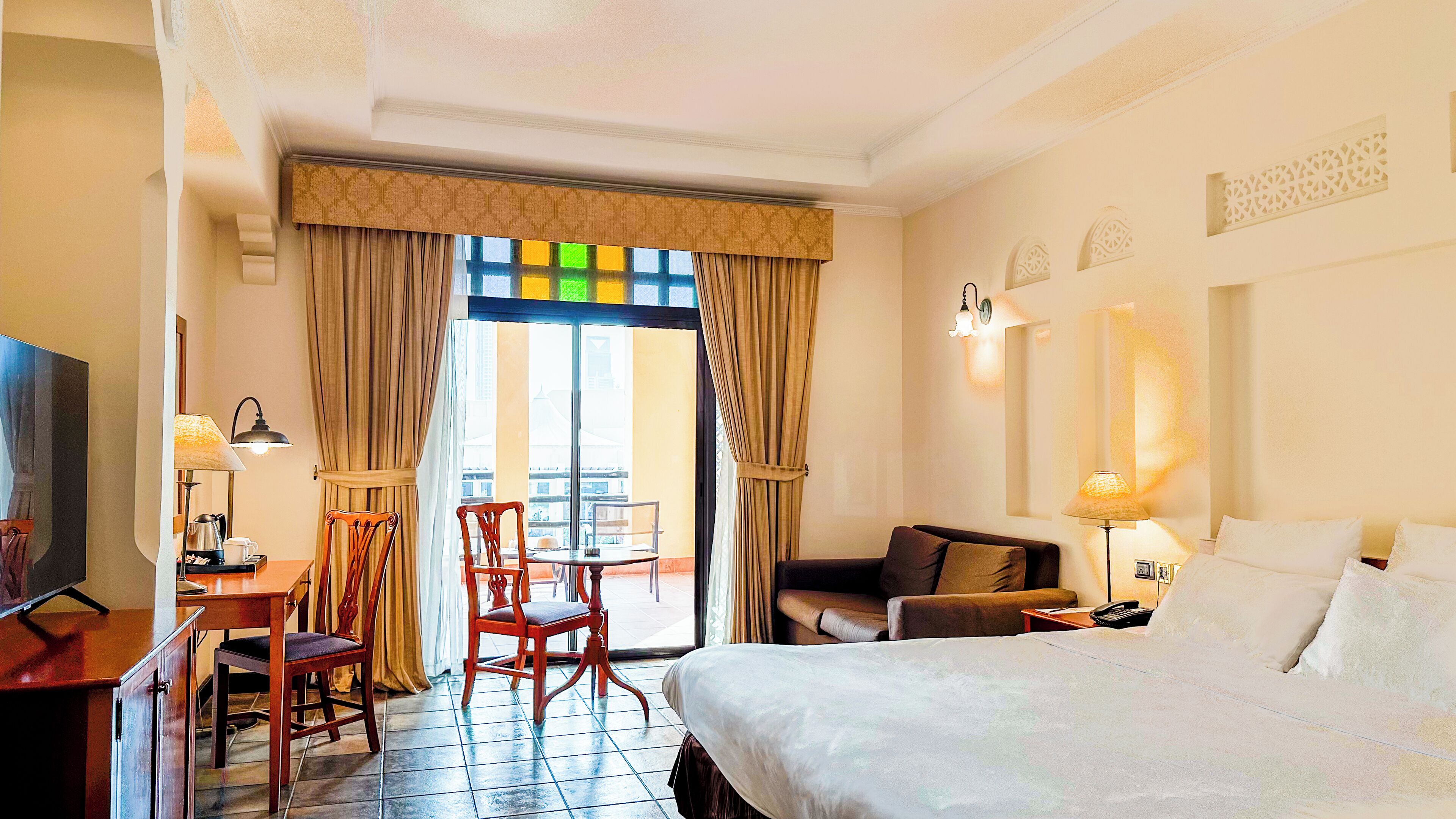 A hotel room with a bed, a desk with a lamp, two chairs, a sofa, and a view of the outside through a large window.