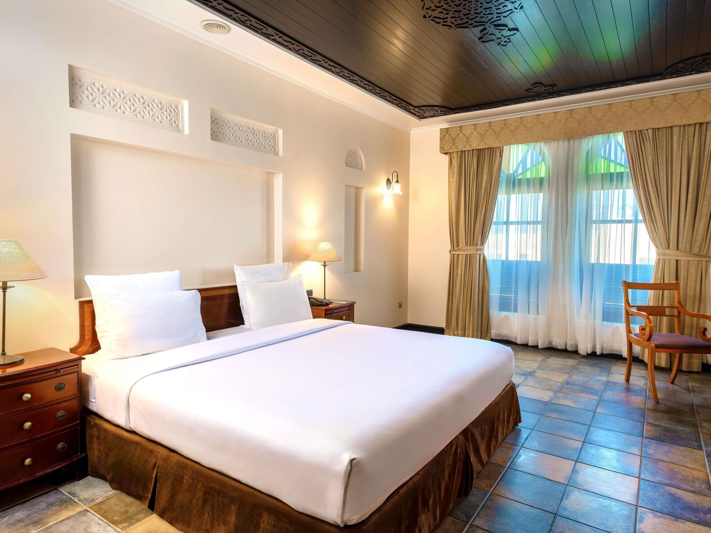A hotel room with a large bed, wooden headboard, a nightstand, a lamp, a chair, and a window with curtains.