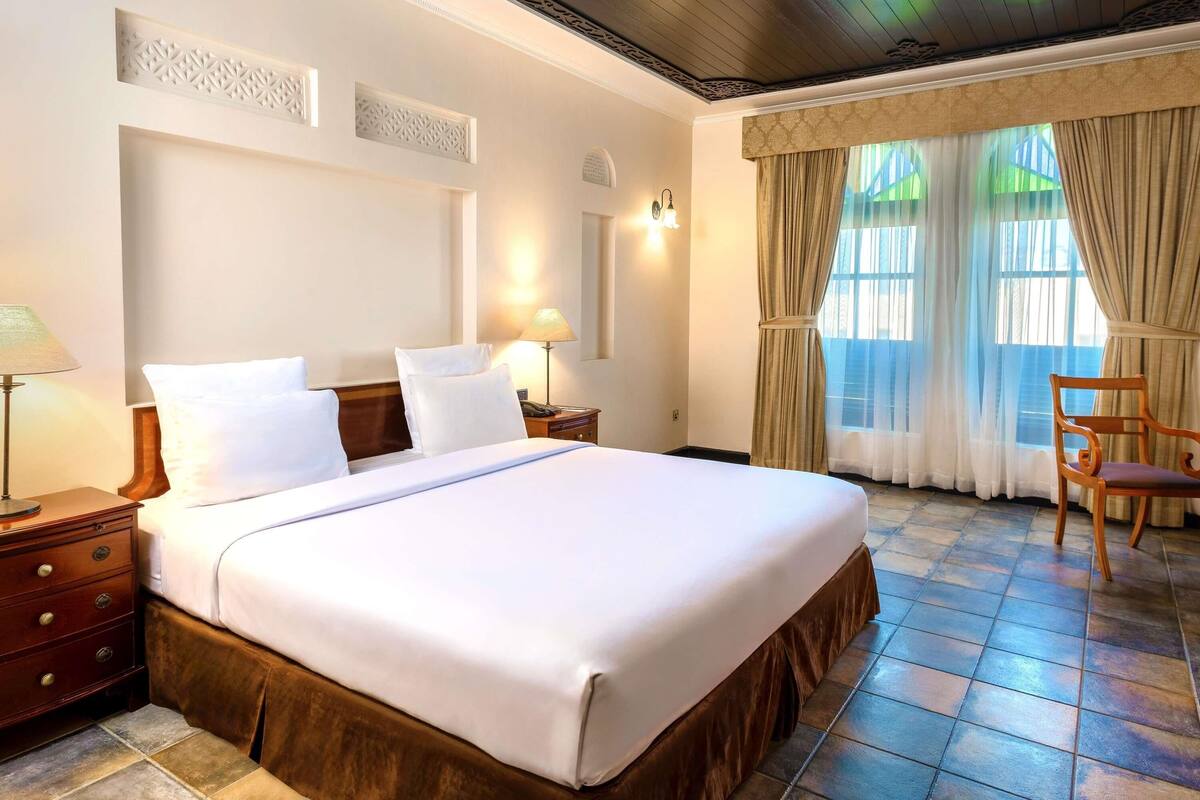A hotel room with a large bed, wooden headboard, a nightstand, a lamp, a chair, and a window with curtains.