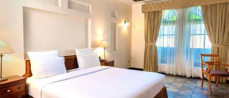 A hotel room with a large bed, wooden headboard, a nightstand, a lamp, a chair, and a window with curtains.