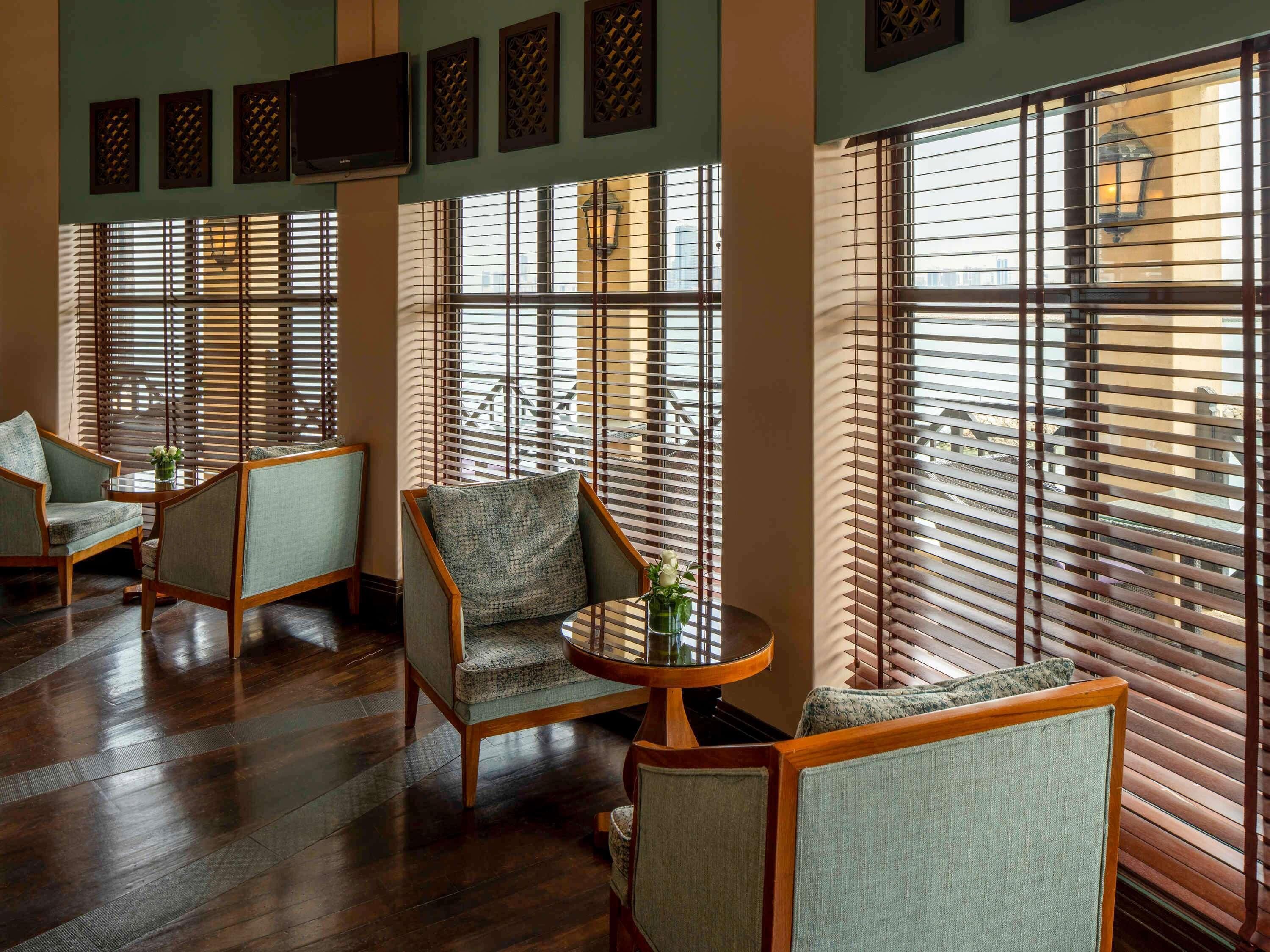 A room with wooden flooring, multiple seating areas with cushioned chairs, and large windows with blinds.