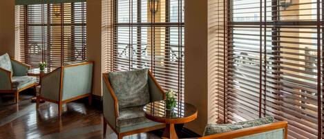 A room with wooden flooring, multiple seating areas with cushioned chairs, and large windows with blinds.