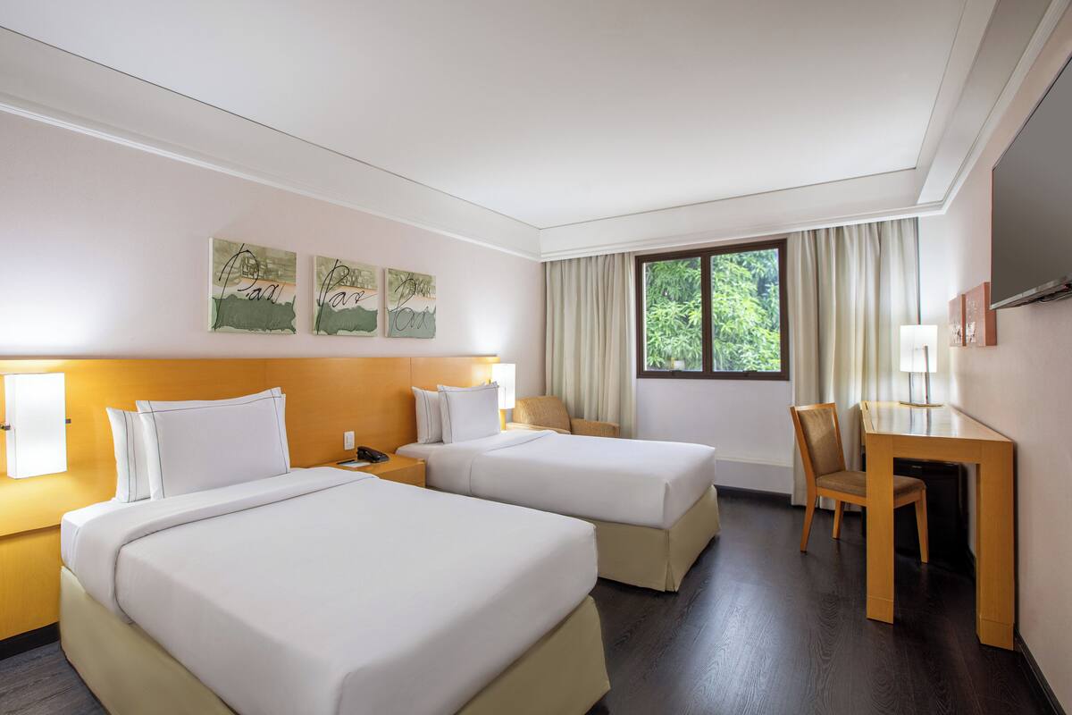 Habitación de hotel con dos camas, un escritorio y una silla. Hay cuadros en las paredes sobre las camas y una ventana con cortinas.