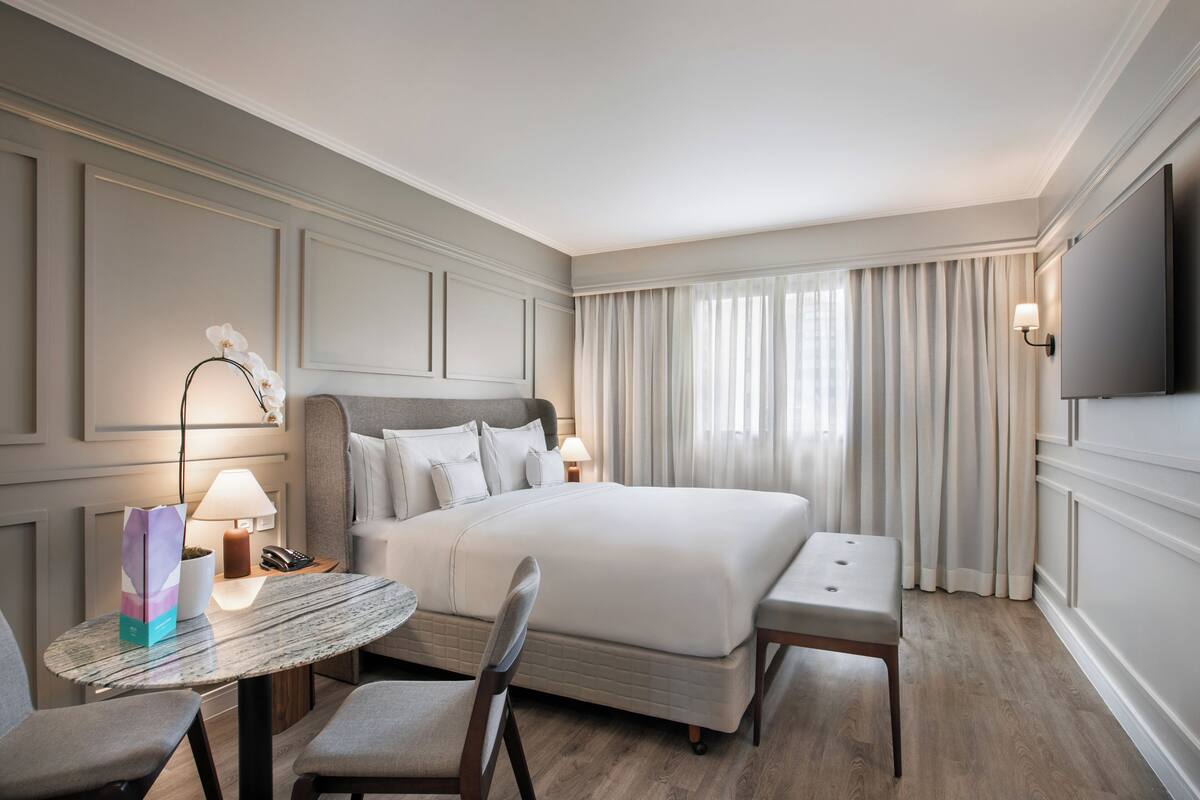 Habitación de hotel con cama, mesitas de noche, escritorio con silla, televisor y ventana con cortinas.