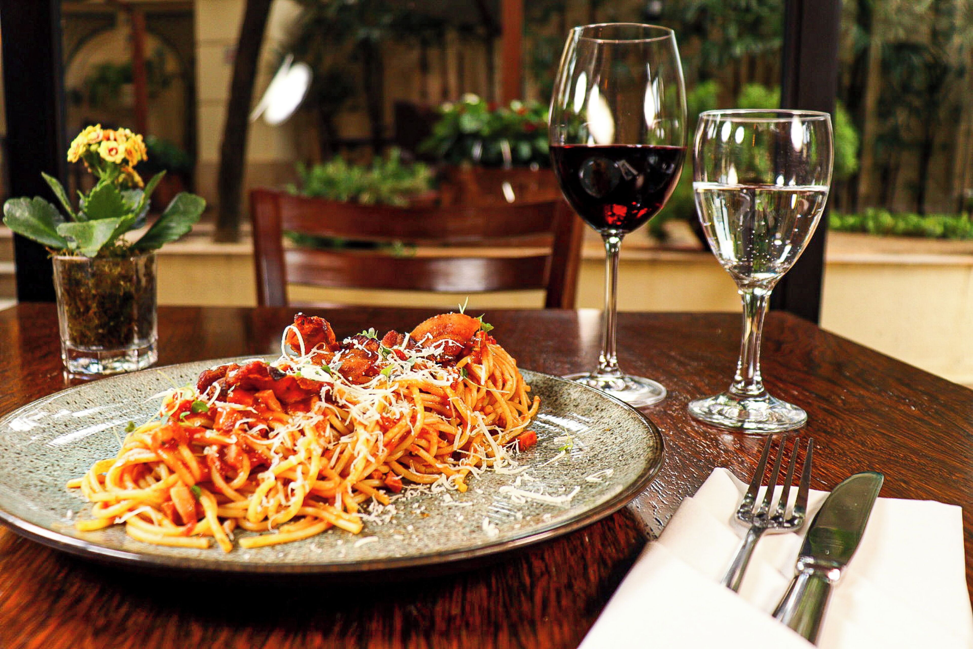 A plate of spaghetti with meat and cheese, two wine glasses, and a small vase with flowers on a wooden table.