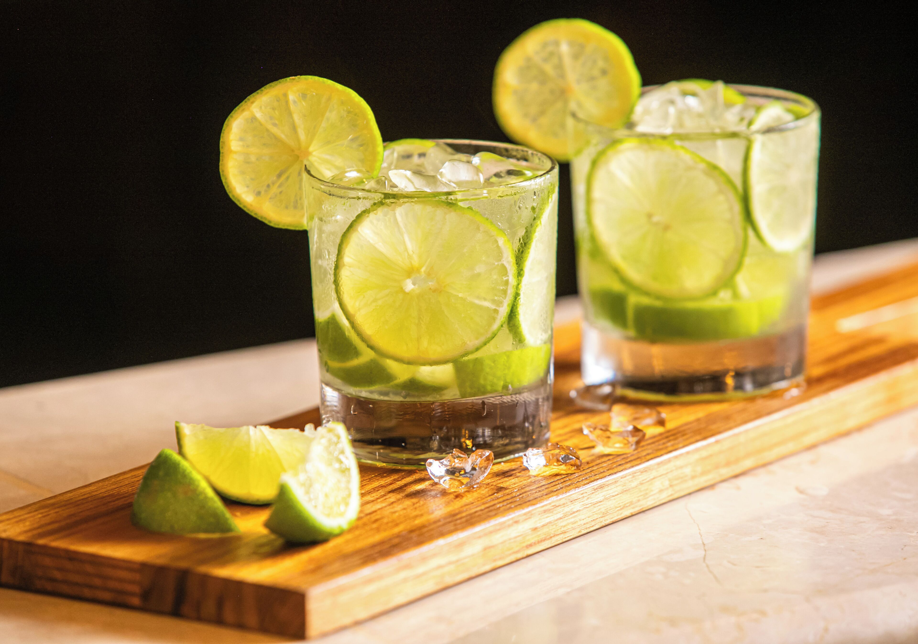 Two glasses of a lime-based beverage with lime slices, ice cubes, and a wooden tray with lime halves.