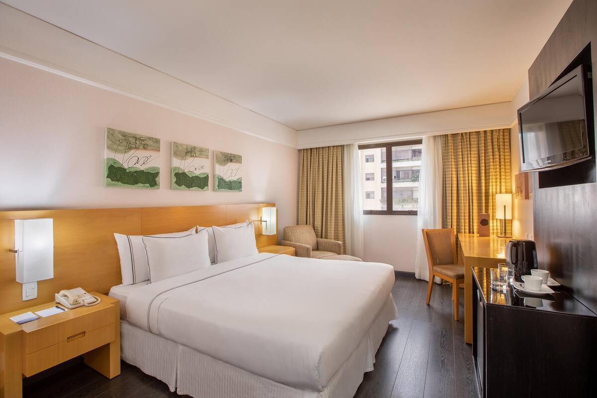 Una habitación de hotel con cama, mesita de noche, lámpara, silla, escritorio, televisor y ventana con cortinas.