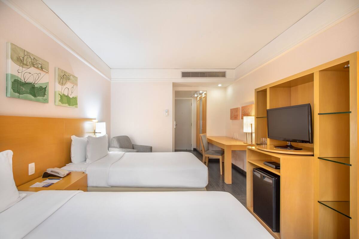 Habitación de hotel con dos camas, un escritorio, televisor, una silla y un refrigerador pequeño.