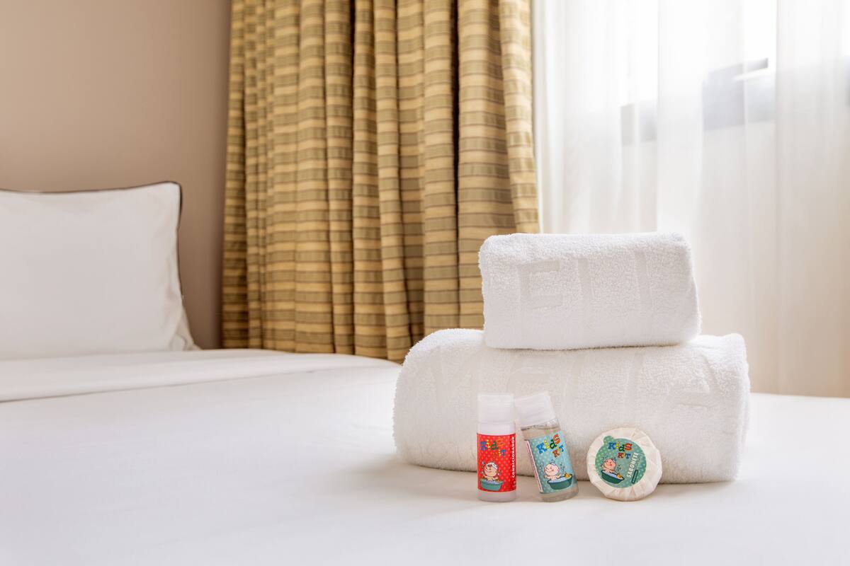 Una cama bien hecha con sábanas blancas, una pila de toallas blancas y dos botellas pequeñas con personajes de dibujos animados en una habitación de hotel.