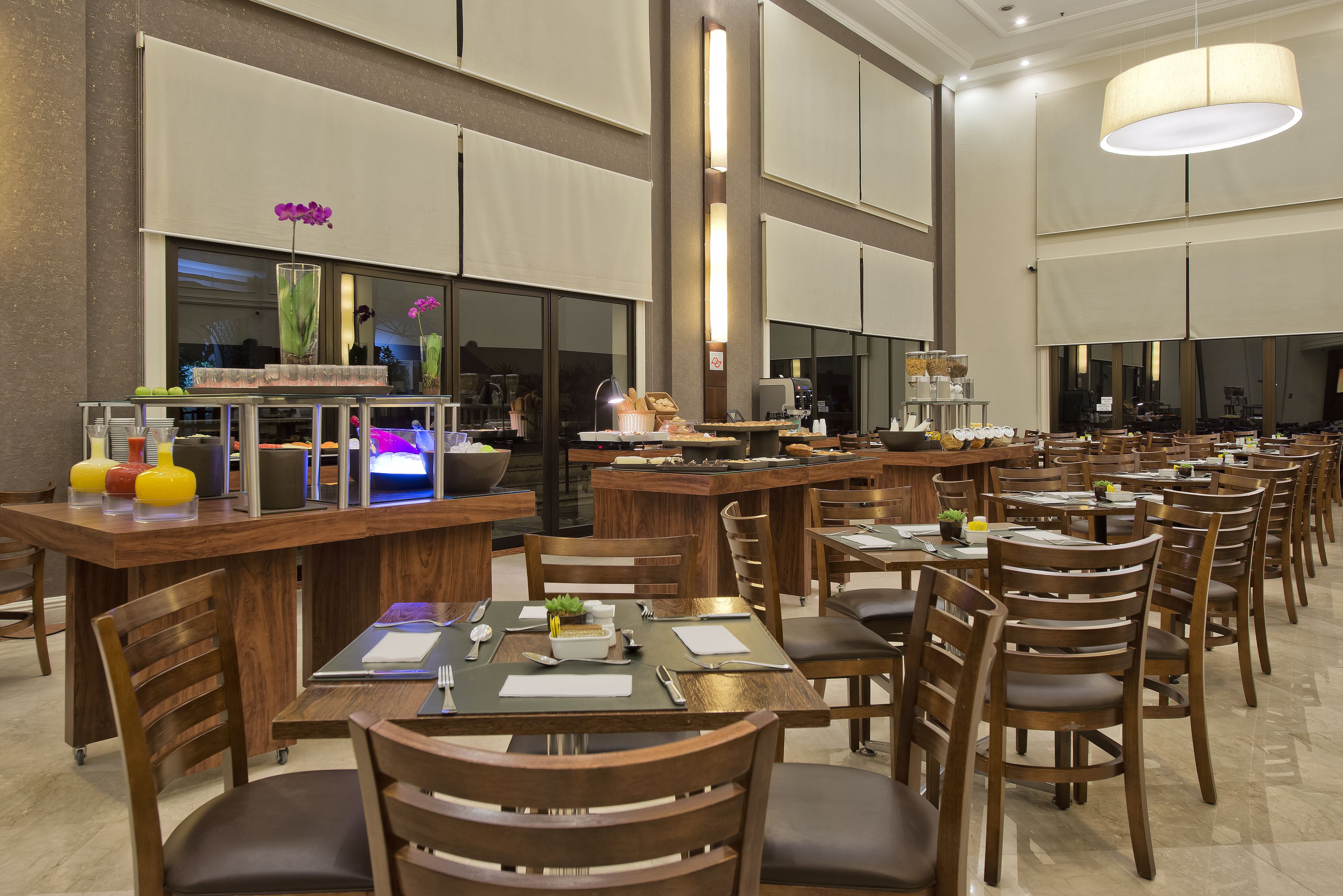 A spacious restaurant with multiple dining tables and chairs, a bar area, and a large window with decorative plants.