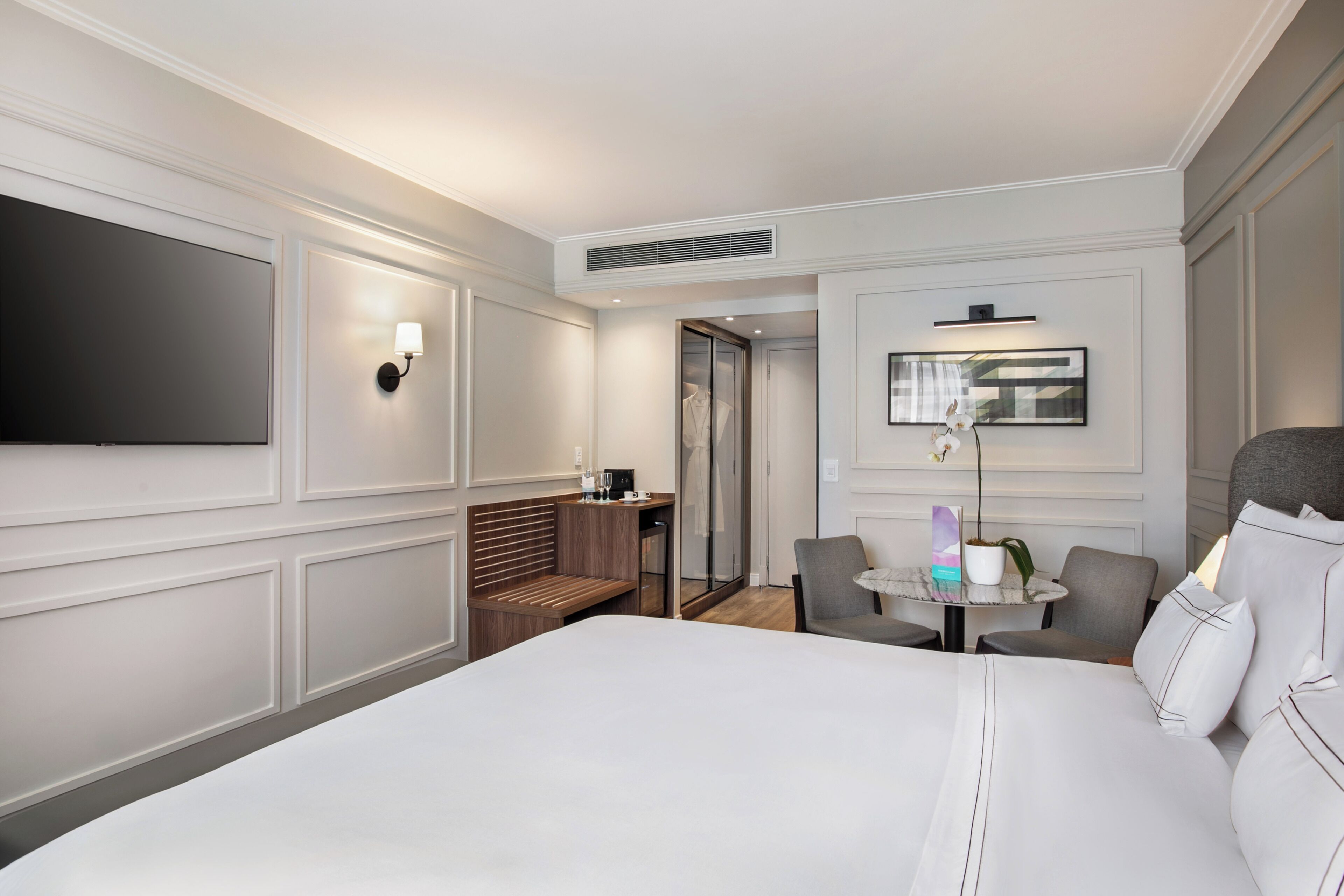 A hotel room with a large bed, a seating area with a table and chairs, a flat-screen TV, and a wall-mounted lamp.
