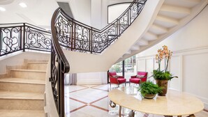 Una imponente escalera con barandales negros ornamentados, piso de mármol y una mesa redonda con plantas en macetas.