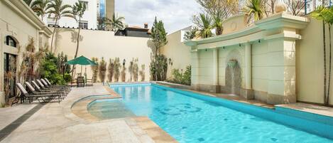 Una piscina con sillas de descanso y una fuente, rodeada de palmeras y la fachada de un edificio.