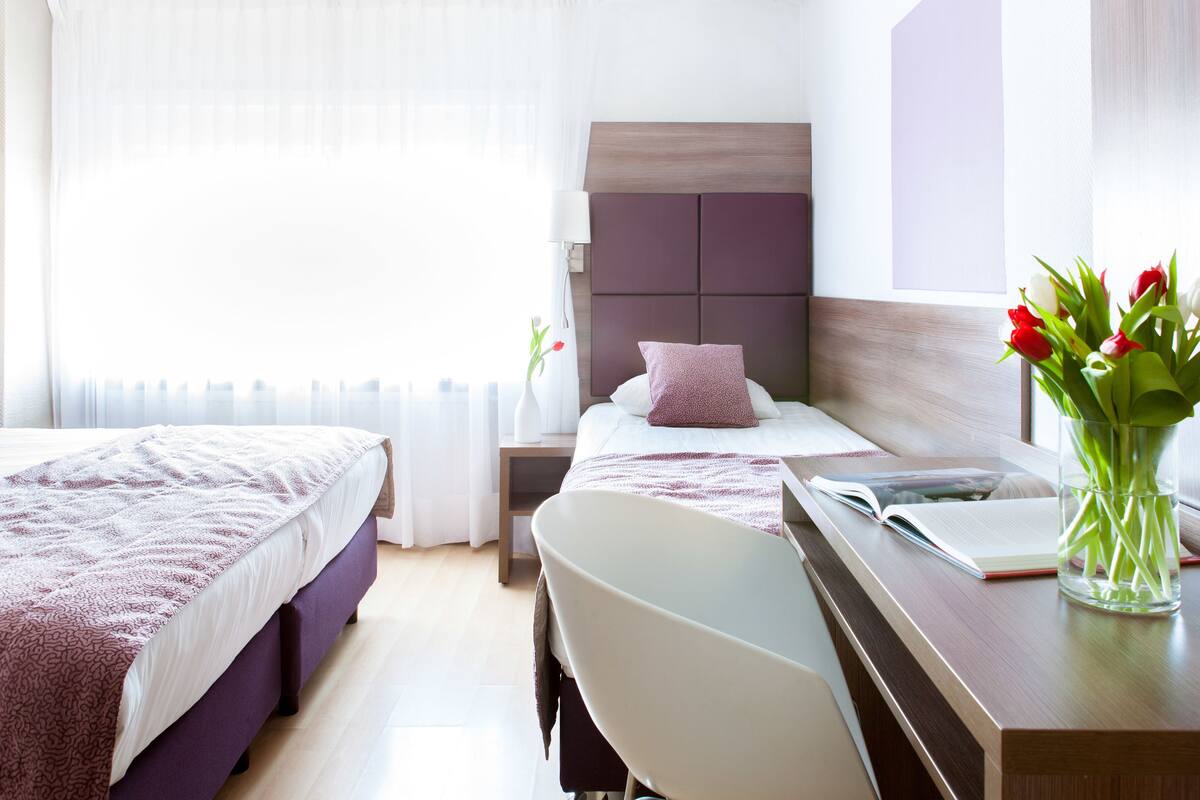 A hotel room with two beds, a desk with a chair, a bedside table with a lamp, and a vase with flowers.