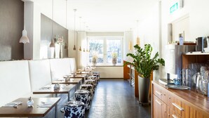 A restaurant with a long counter, multiple tables, and a row of stools. There are hanging lights, a large window, and a potted plant. - Hotel Victorie (Amsterdam)