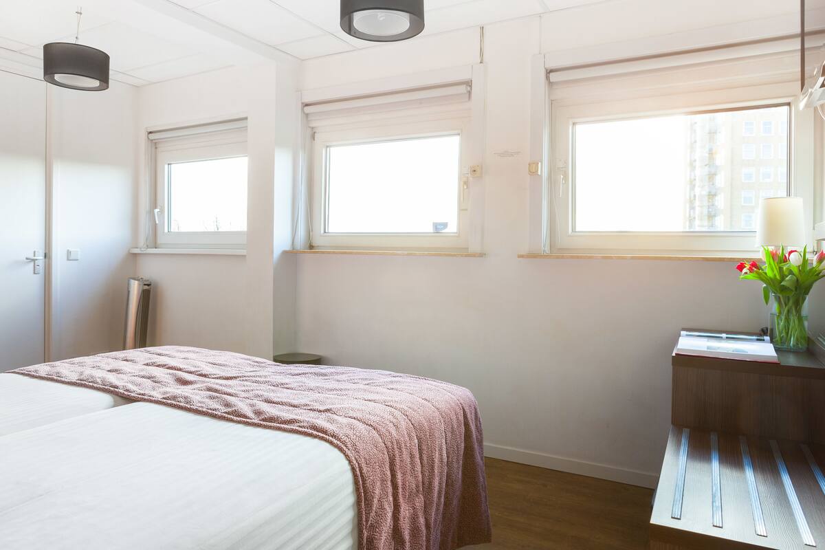 A bedroom with a bed, a bedside table with a lamp, and a window with a view of buildings.