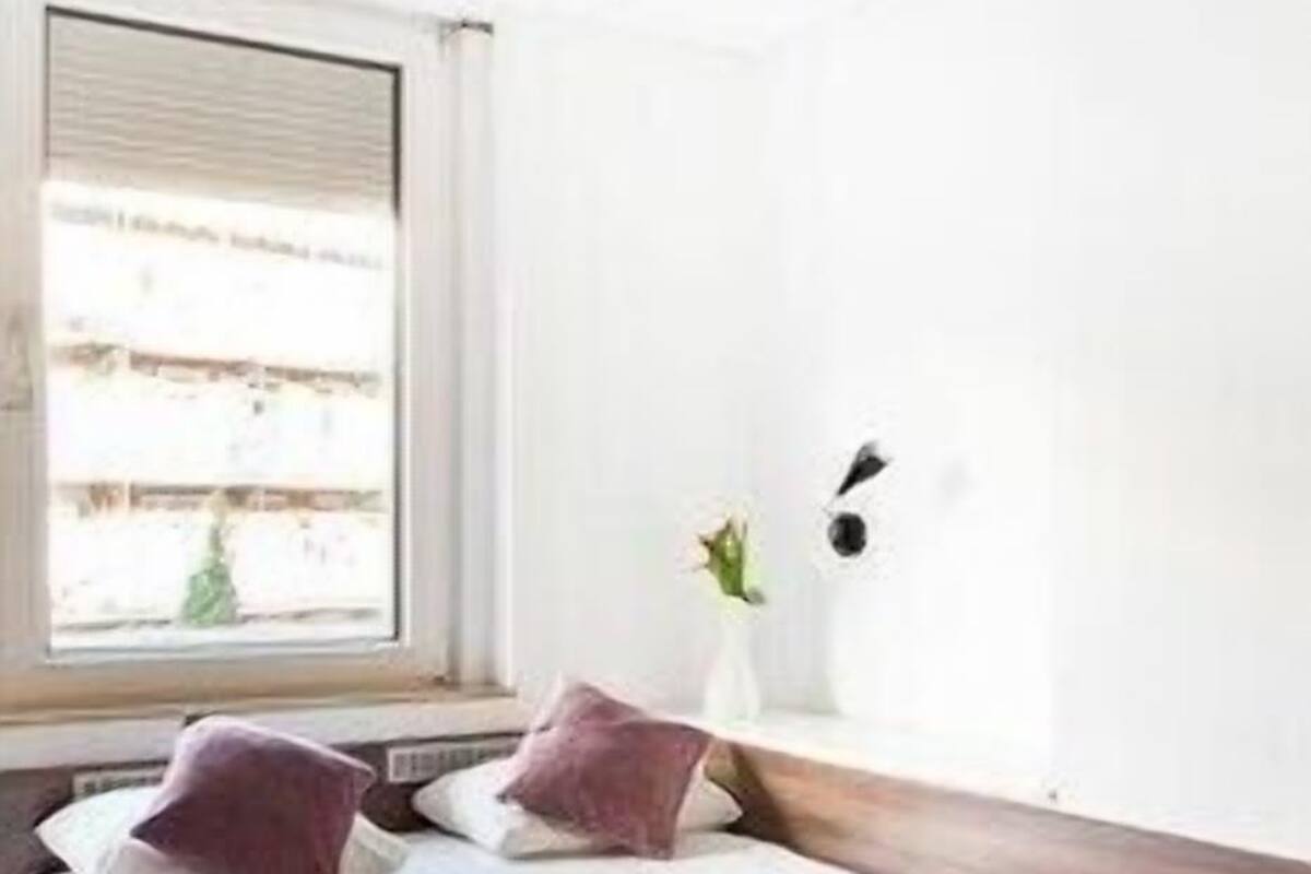 A bedroom with a bed, a window, a wall-mounted light fixture, and a bedside table.