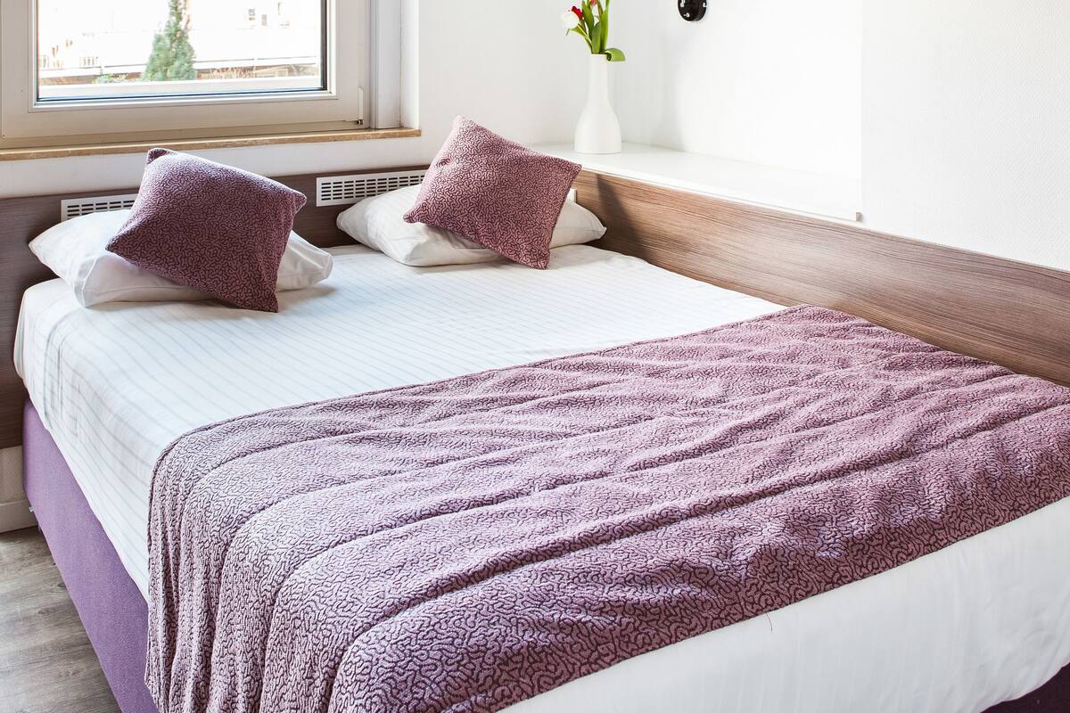 A modern bedroom with a large bed, a bedside table with a vase, and a window with blinds.