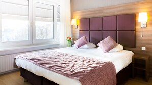 A hotel room with a large bed, a window with blinds, a bedside table with a lamp, and a wall-mounted light fixture. - Hotel Victorie (Amsterdam)