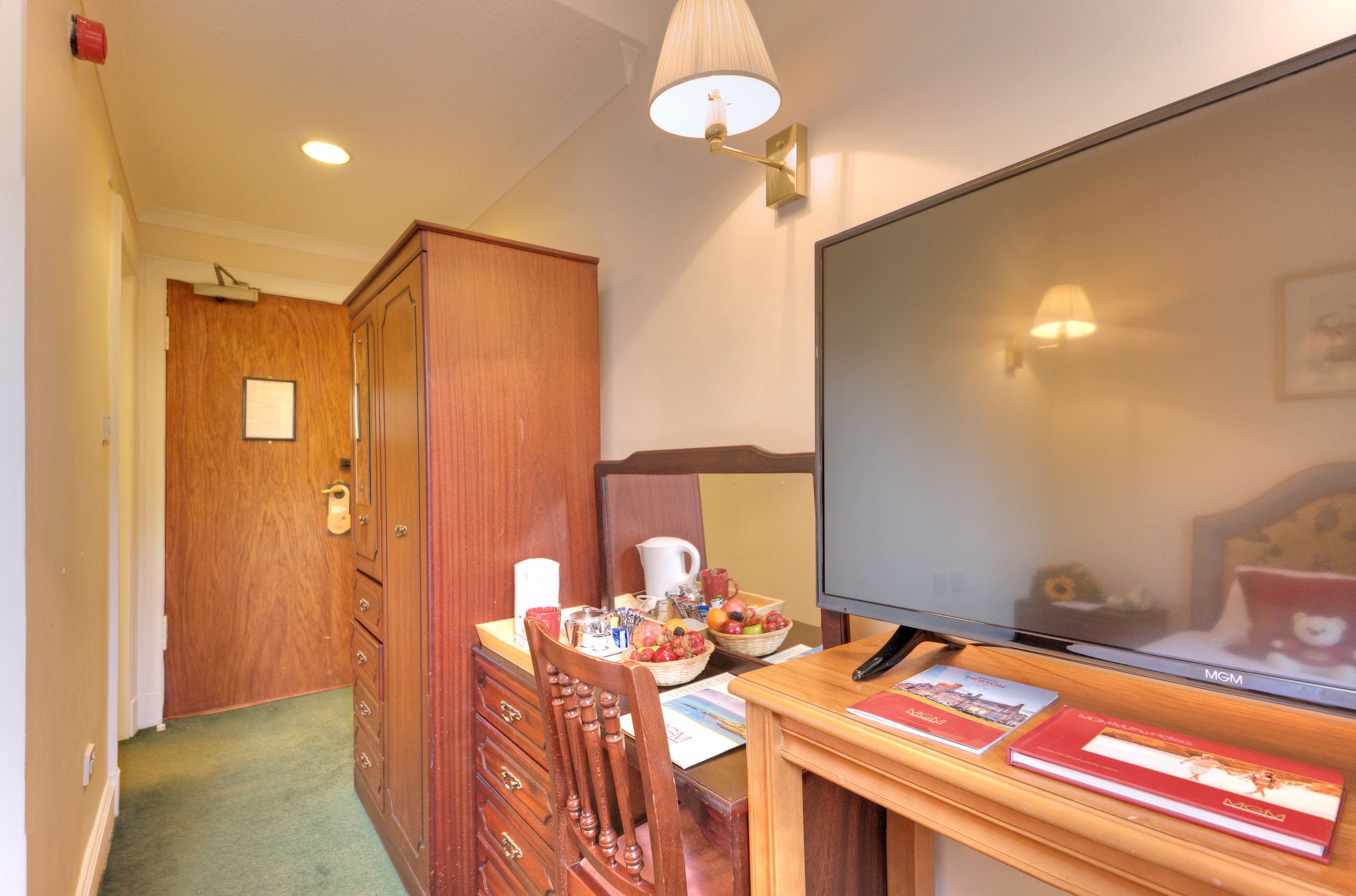 A hotel room with a wooden desk, a television, a wooden wardrobe, and a door leading to another room.