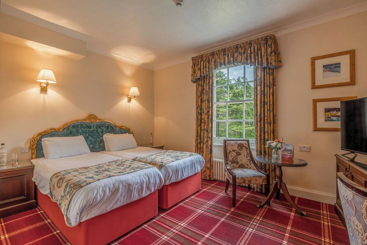 A hotel room with a double bed, two bedside lamps, a television, a small table with a vase, and a window with curtains.