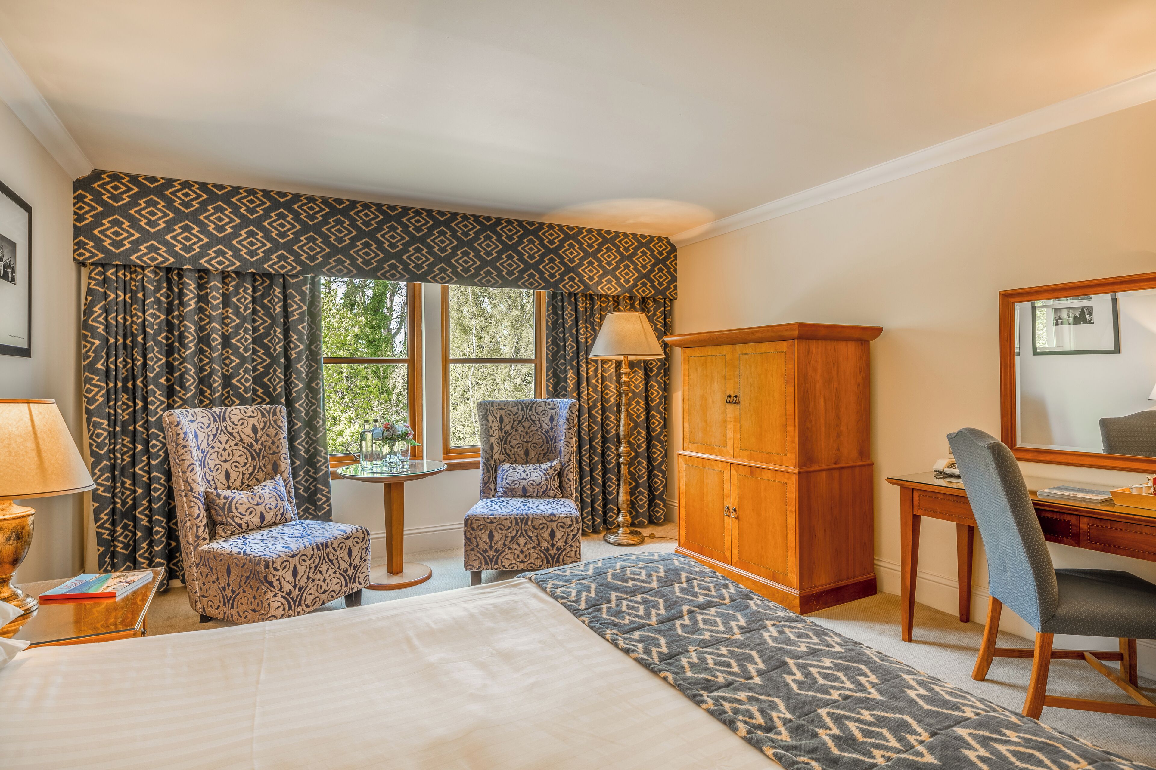 Habitación de hotel con cama, dos sillones estampados, un aparador de madera, un escritorio con silla y una ventana con cortinas.