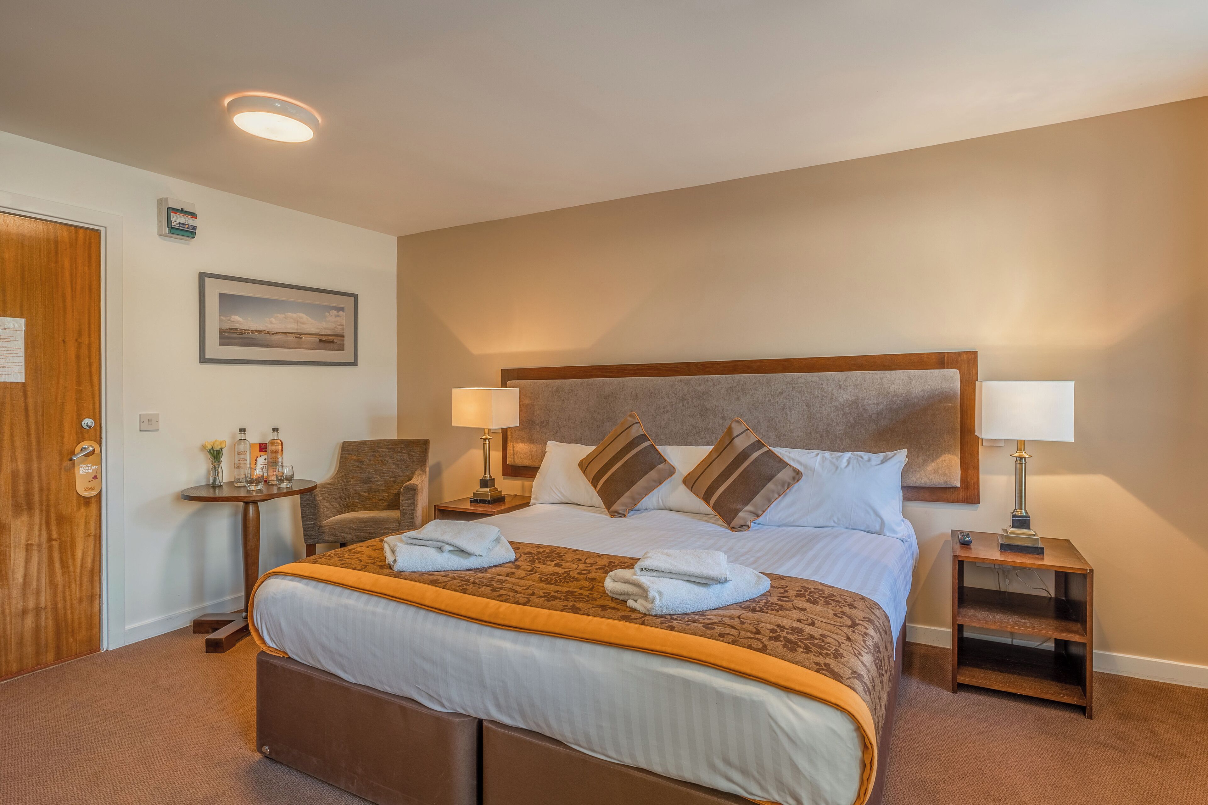 A hotel room with a large bed, two bedside tables, a chair, and a small table with a lamp.