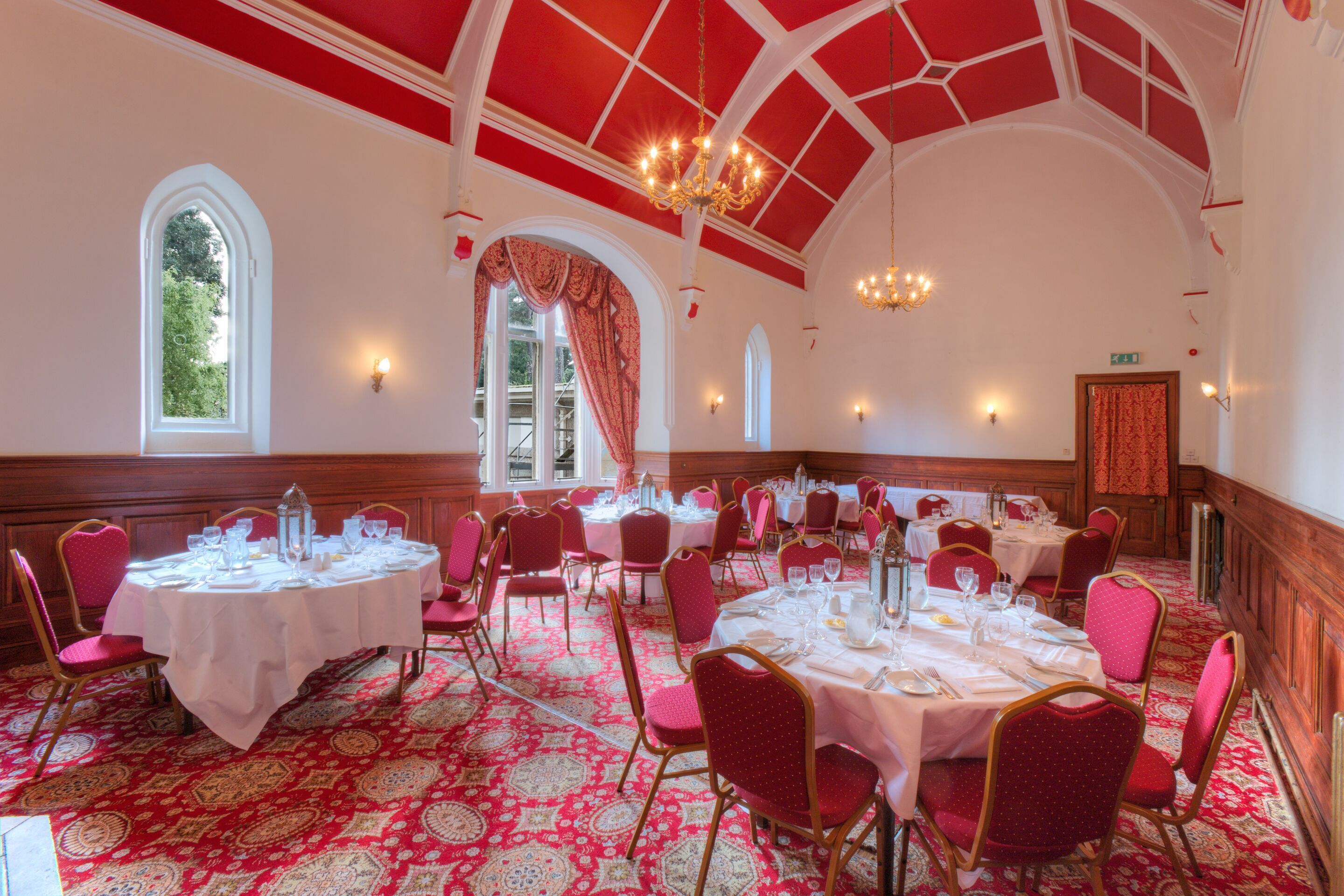 A banquet hall with round tables, red chairs, and chandeliers.