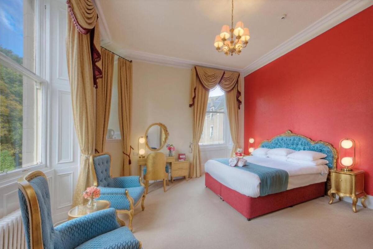 A bedroom with a red wall, a large bed with a blue headboard, a chandelier, and a view of greenery outside.