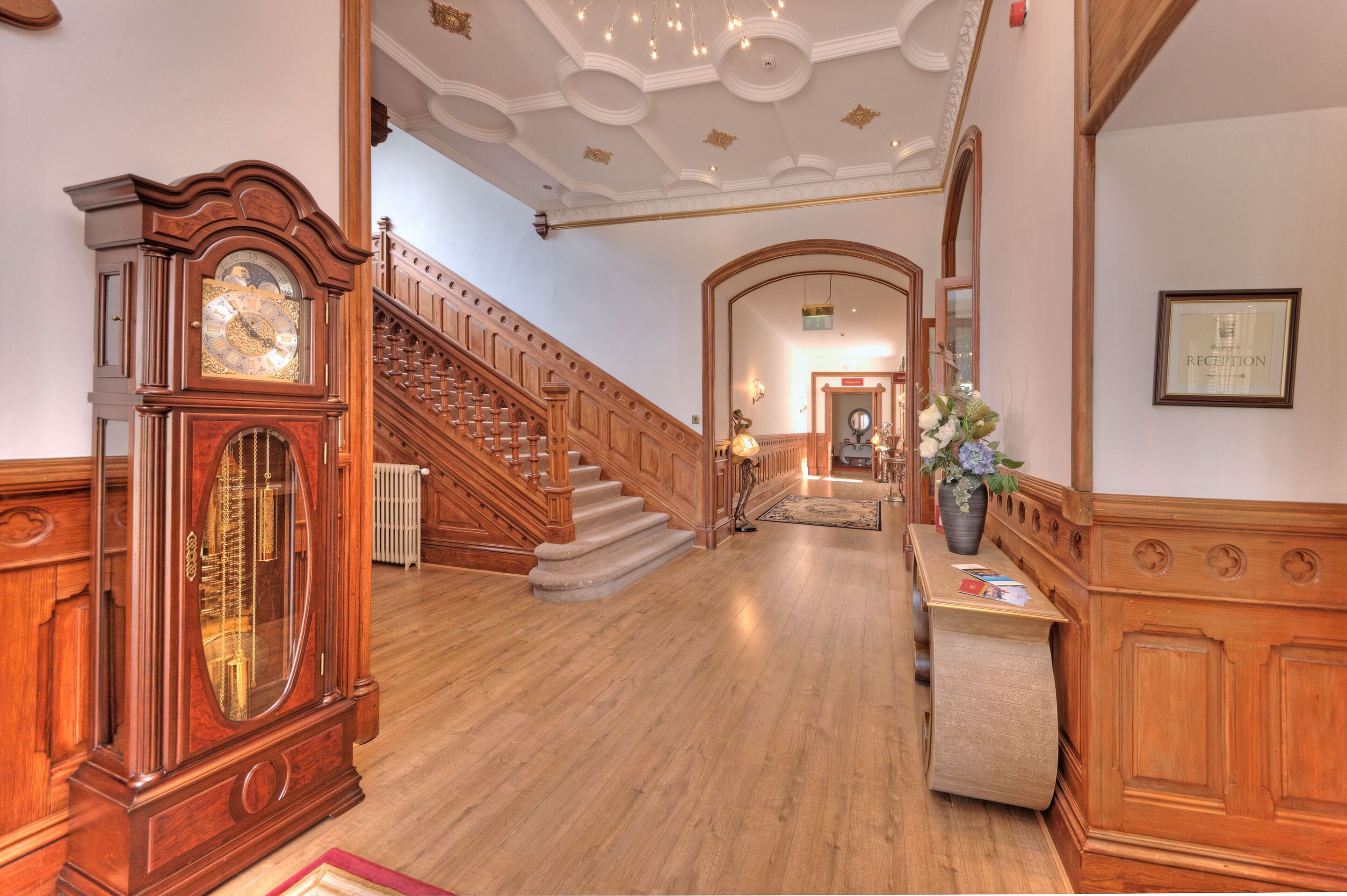 A grand hallway with a wooden staircase, a large grandfather clock, and a reception desk with a vase of flowers.