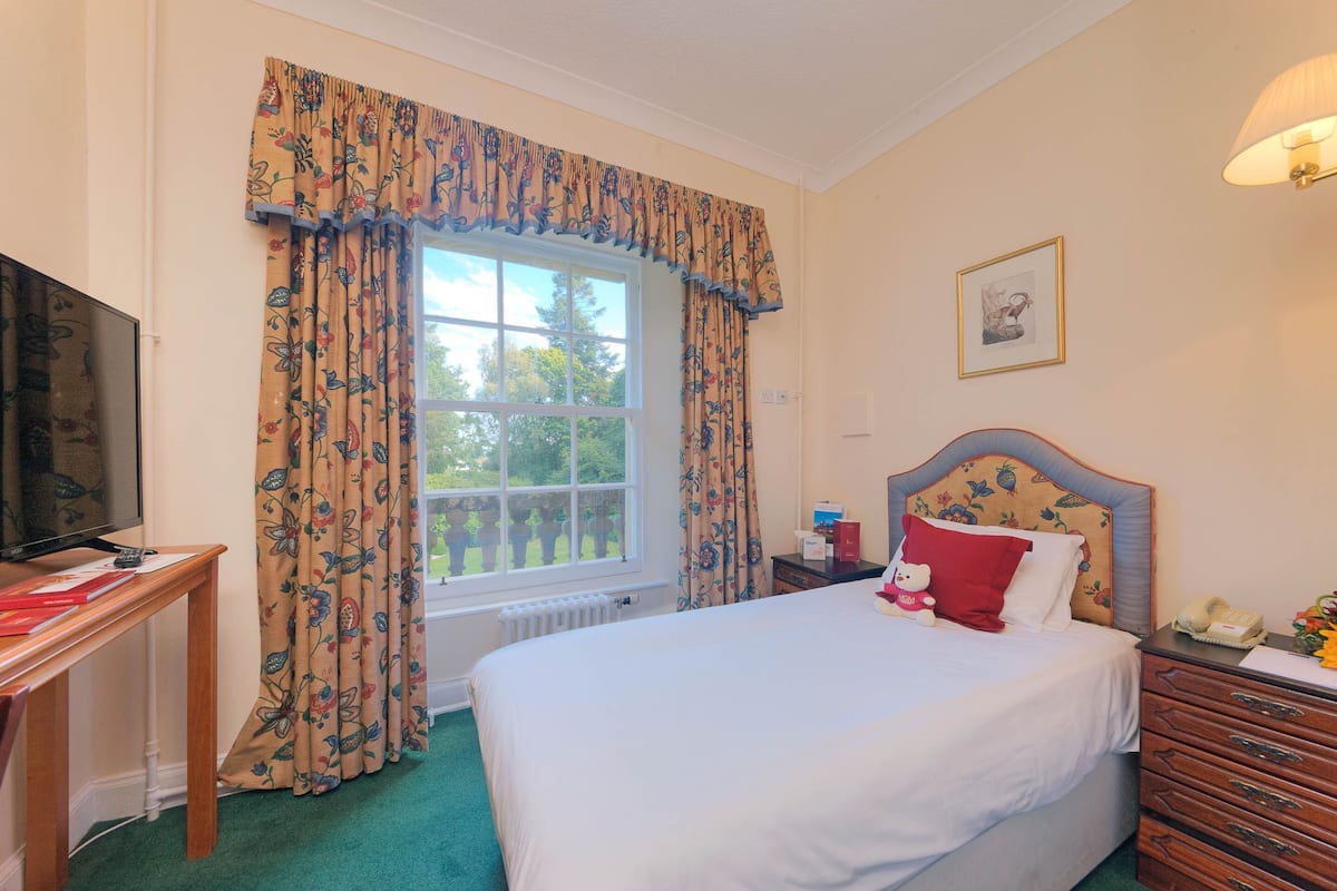 A bedroom with a bed, a bedside table, a lamp, a window with curtains, a television, and a framed picture on the wall.