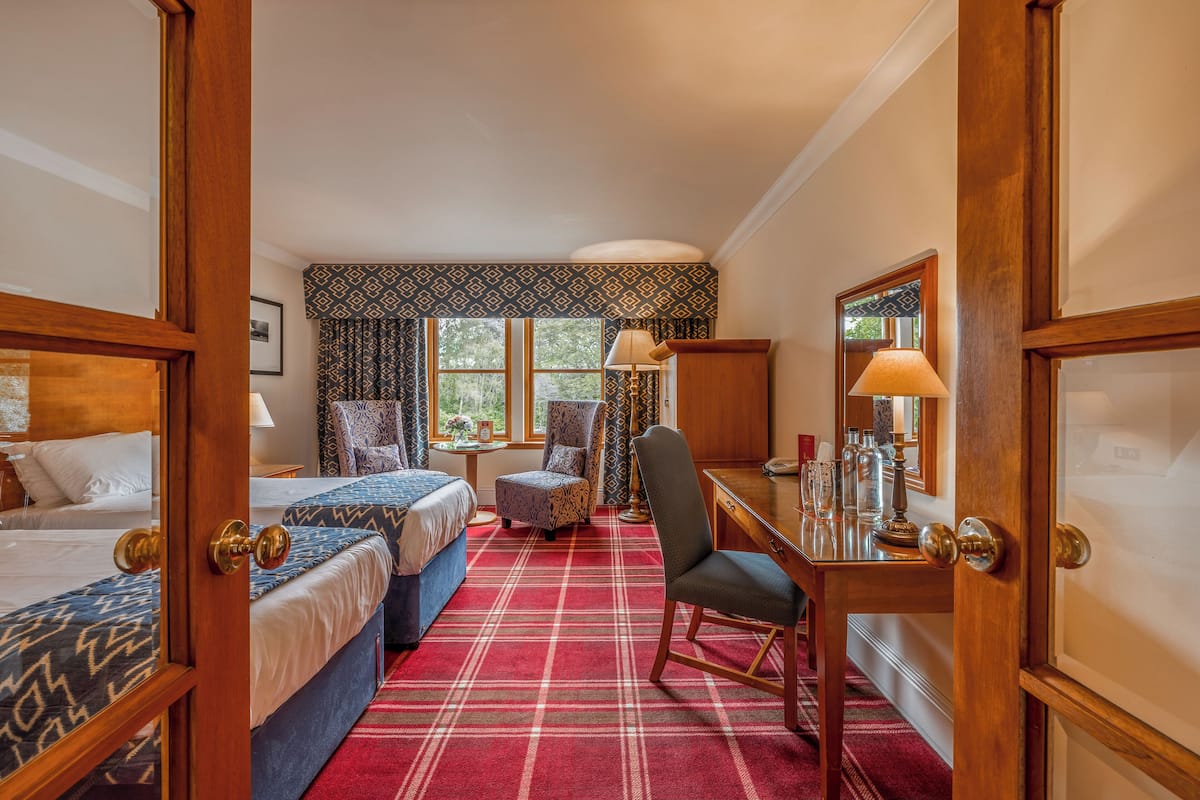A hotel room with two beds, a desk with a lamp, a chair, and a window with curtains.