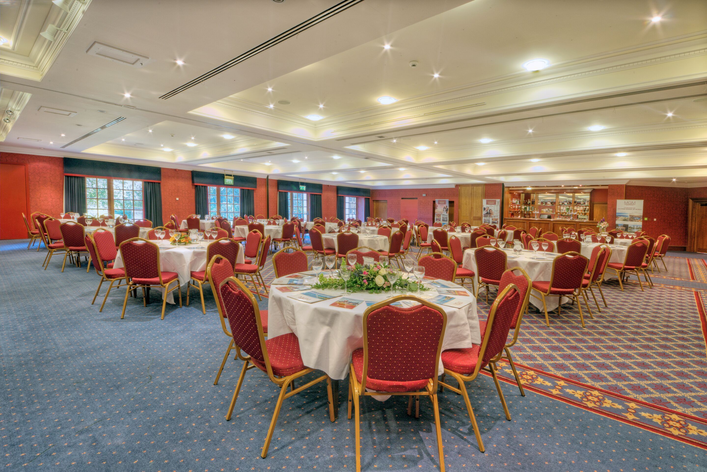 A large banquet hall with round tables and red chairs, carpeted floor, and a bar area in the background.