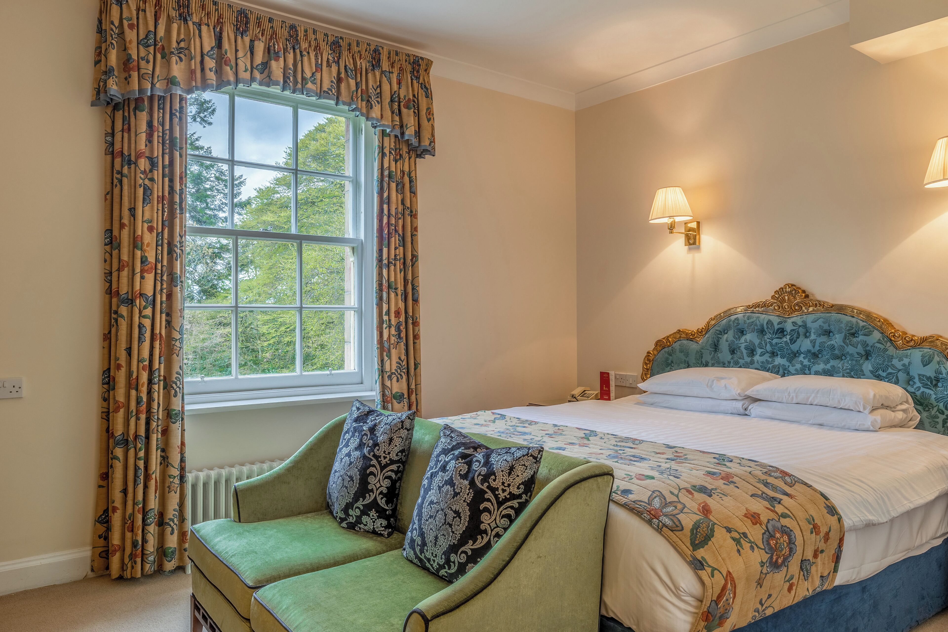 Un dormitorio con sofá verde, cama con ropa de cama floral, ventana con cortinas y luces fijadas a la pared.