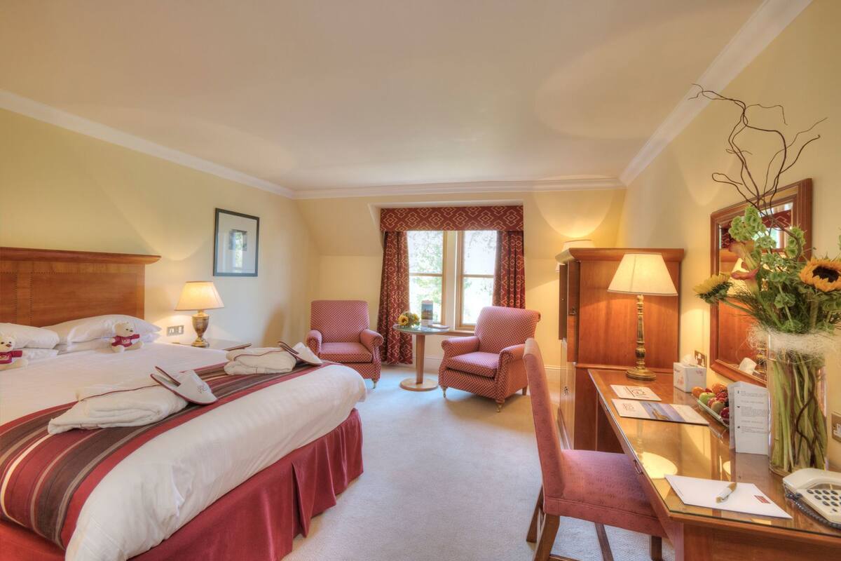 A hotel room with a bed, two chairs, a desk, a lamp, a vase with flowers, and a mirror.