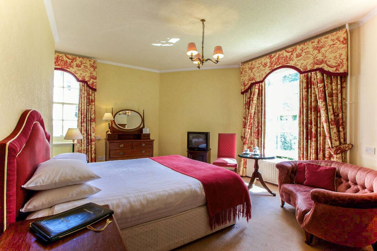 A hotel room with a large bed, a red armchair, a small table with a lamp, and a television.