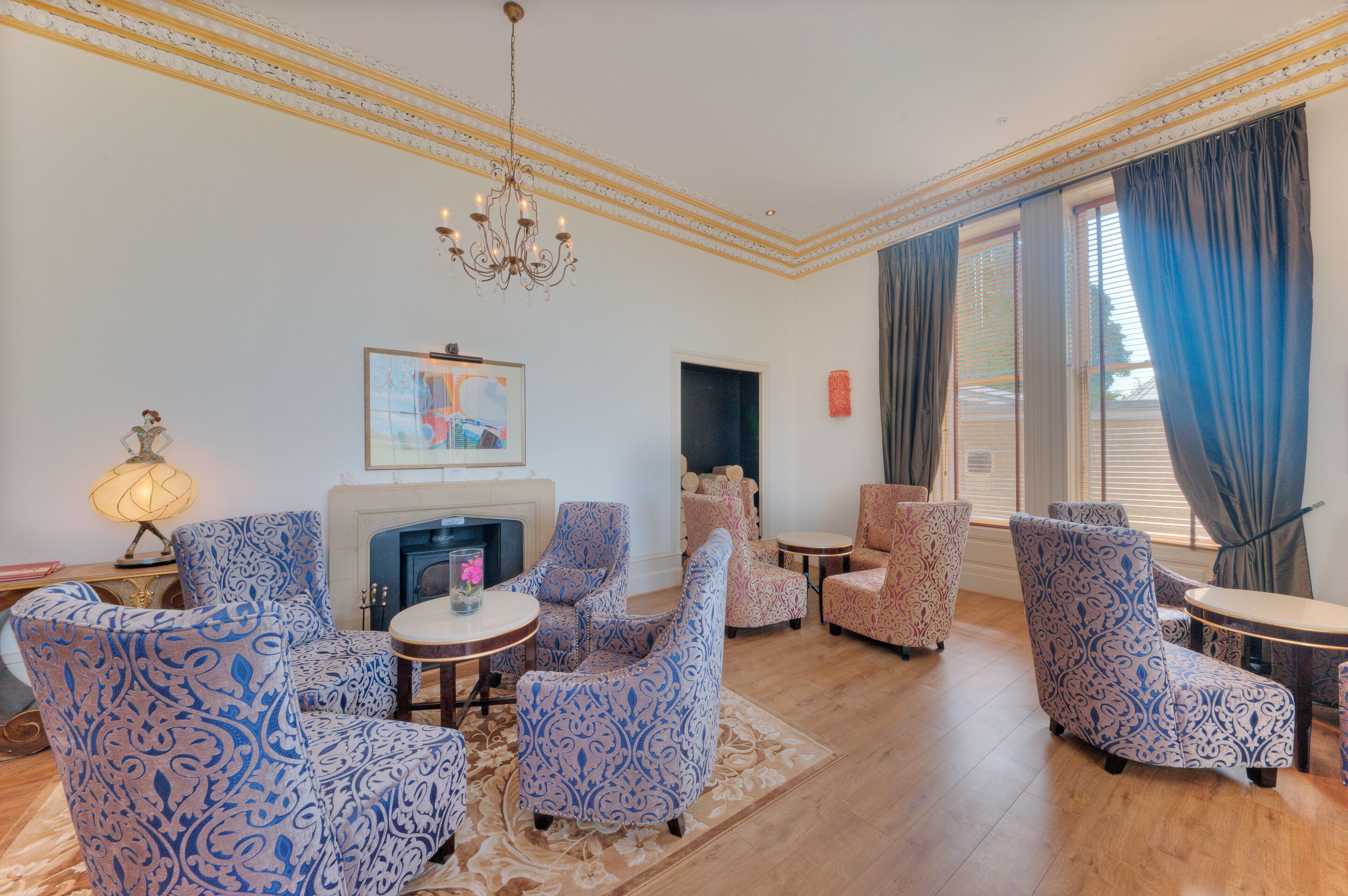 A living room with patterned blue and pink chairs, a chandelier, a fireplace, and a large window with curtains.