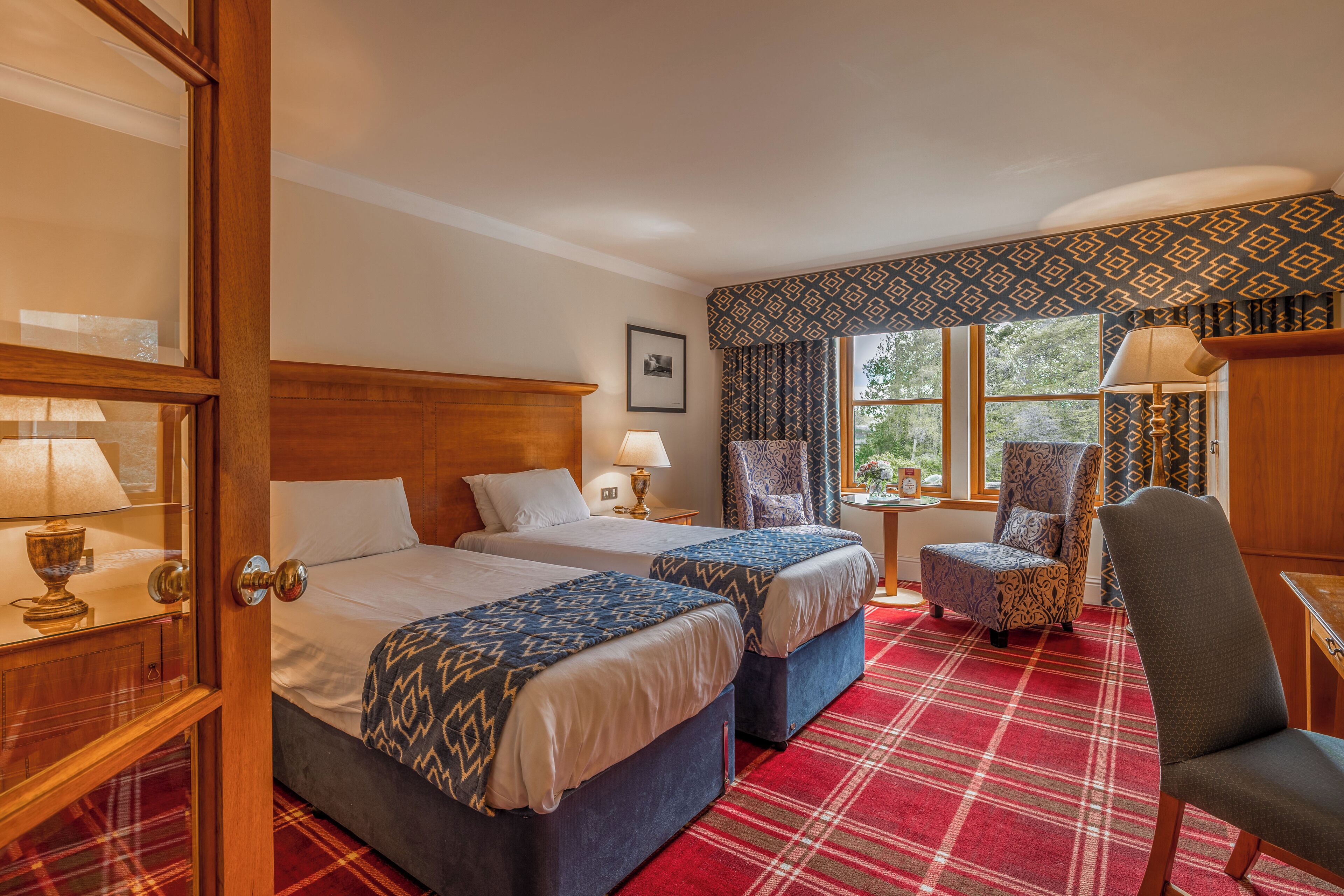 A hotel room with two beds, a red plaid carpet, a wooden wardrobe, a chair, and a window with patterned curtains.