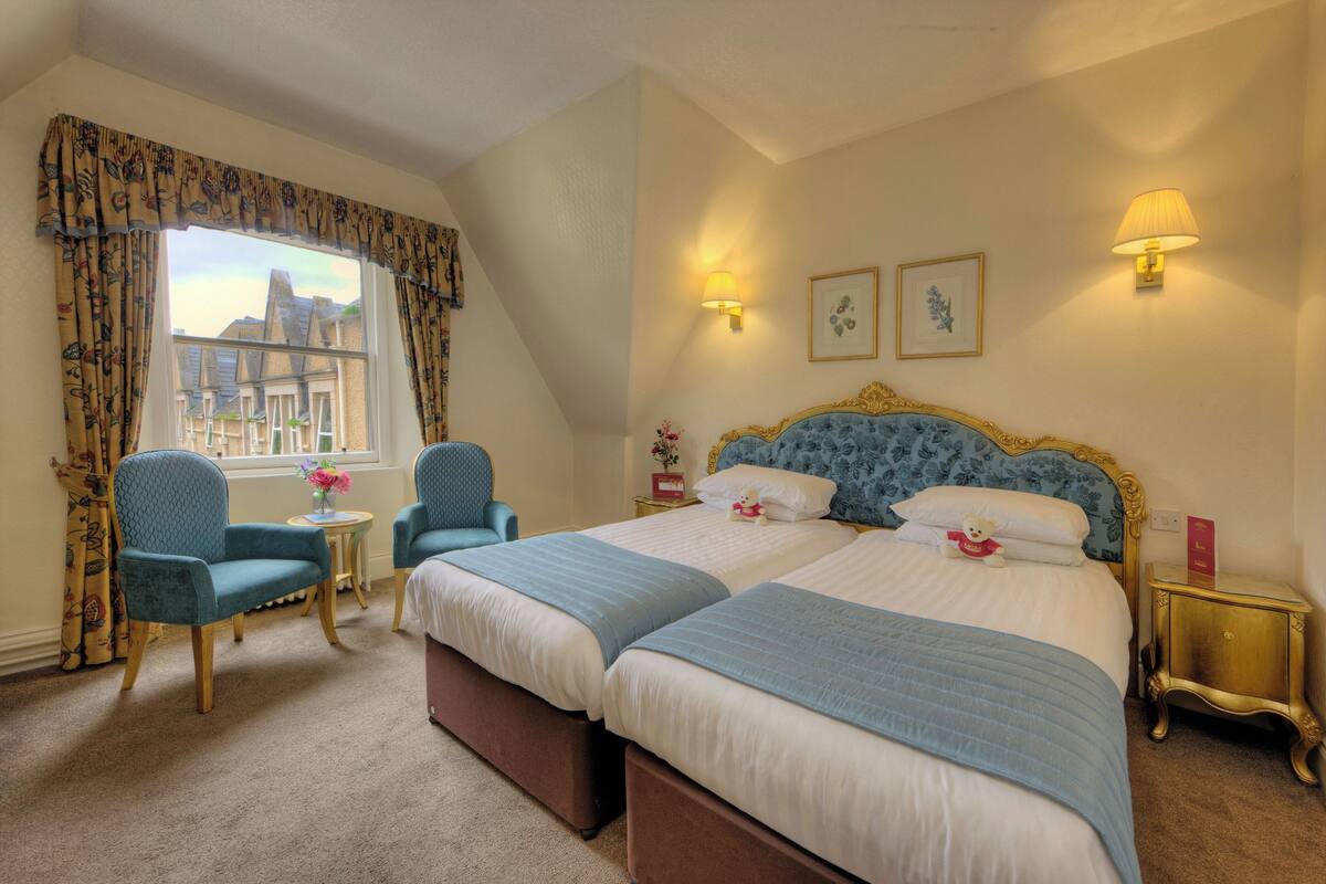 A hotel room with two beds, a sitting area with chairs, a small table with a vase of flowers, and a window with curtains.