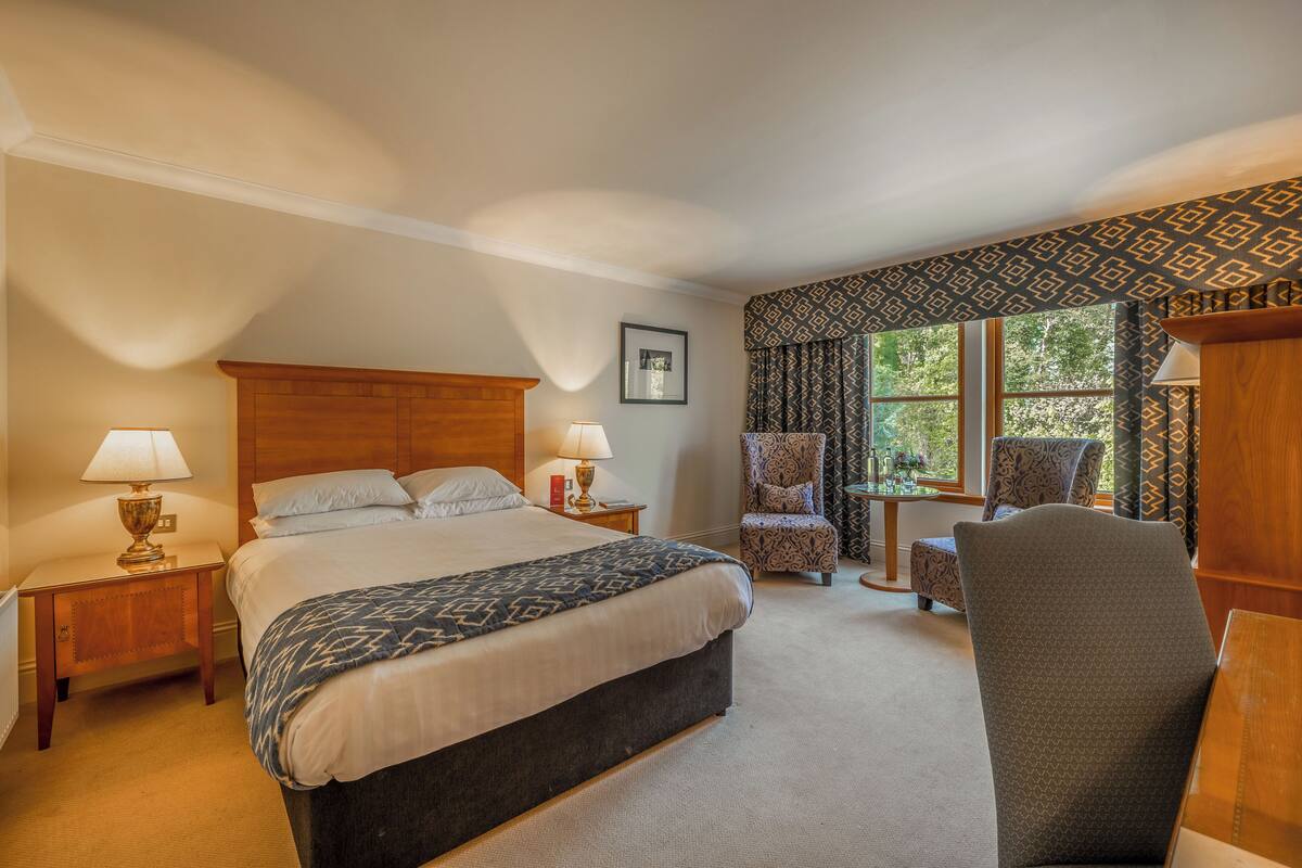 A hotel room with a large bed, two bedside tables with lamps, a small table with a chair, and a window with curtains.