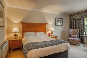A hotel room with a large bed, two bedside tables with lamps, a chair, and a window with patterned curtains.