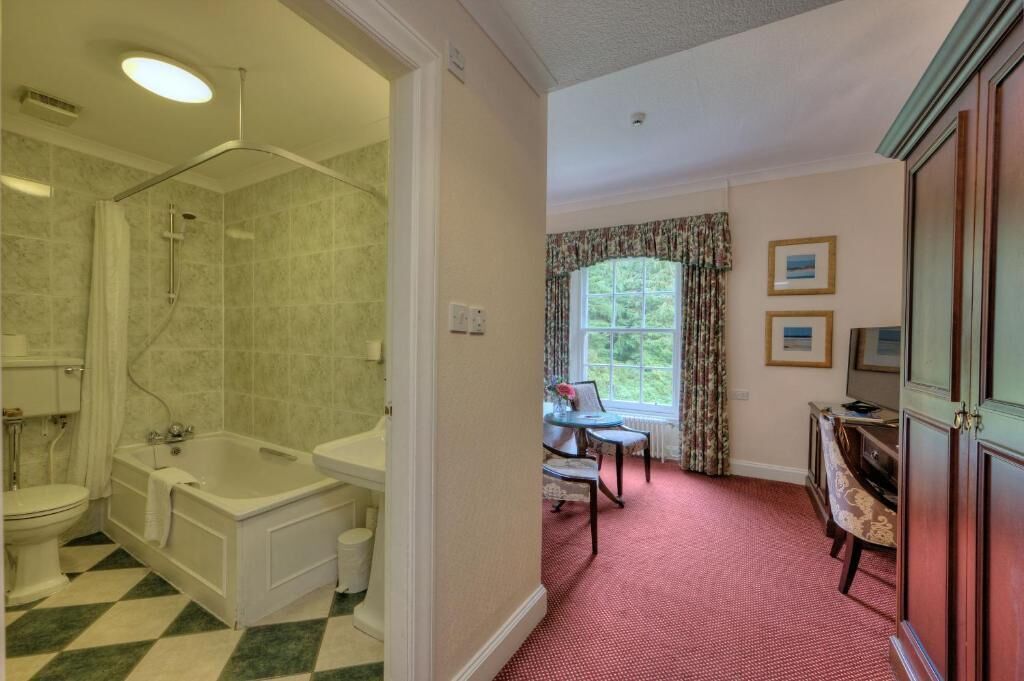 A bathroom with a bathtub, toilet, and shower. There is a window with curtains, a chair, and a small table with a lamp.