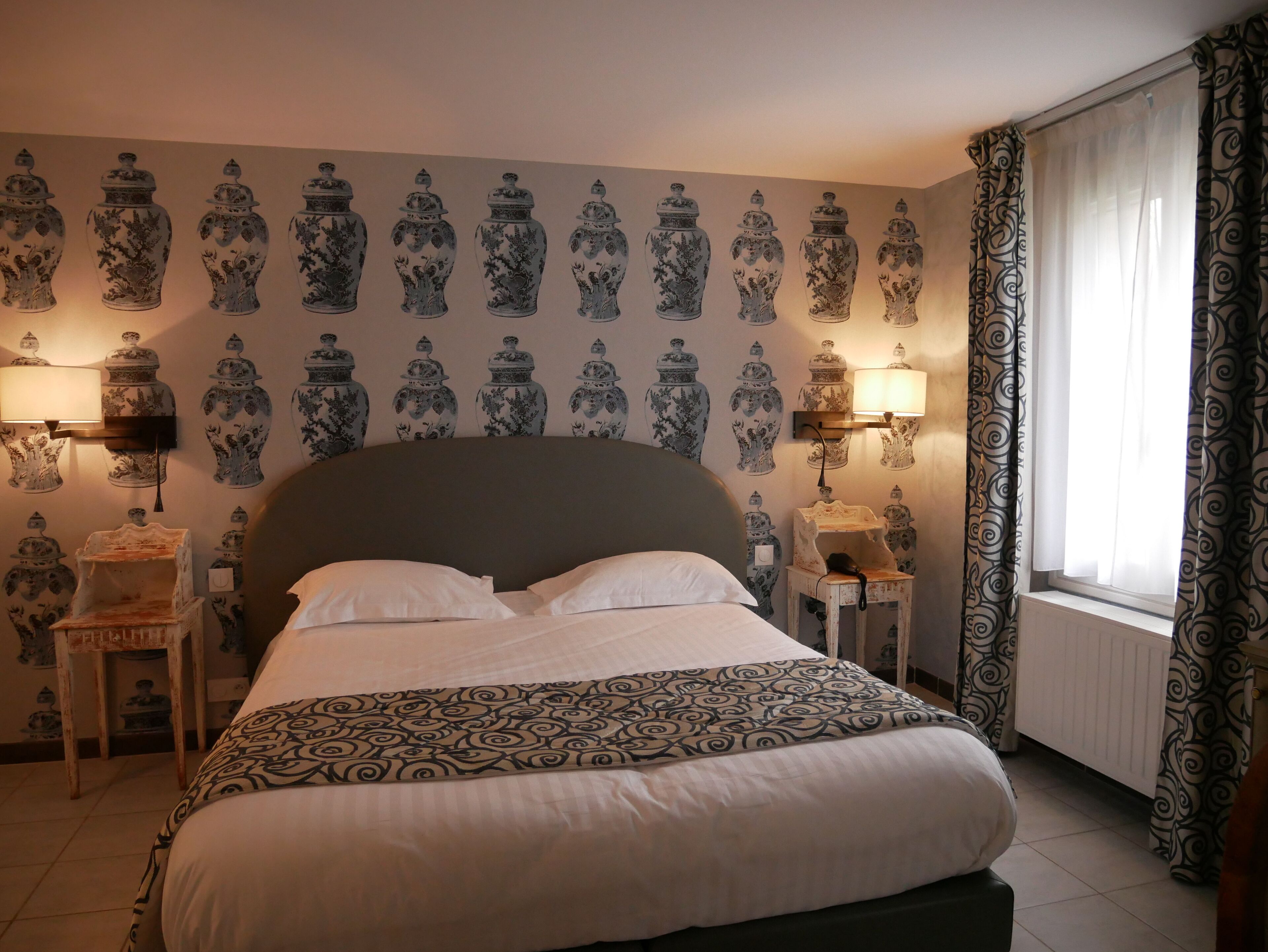 A bedroom with a large bed, two bedside tables with lamps, a patterned bedspread, and a wall with a repeating pattern of vases.