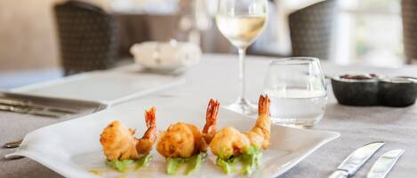 Una mesa de comedor con un plato de entrantes de camarones, una copa de vino blanco y un recipiente con agua.