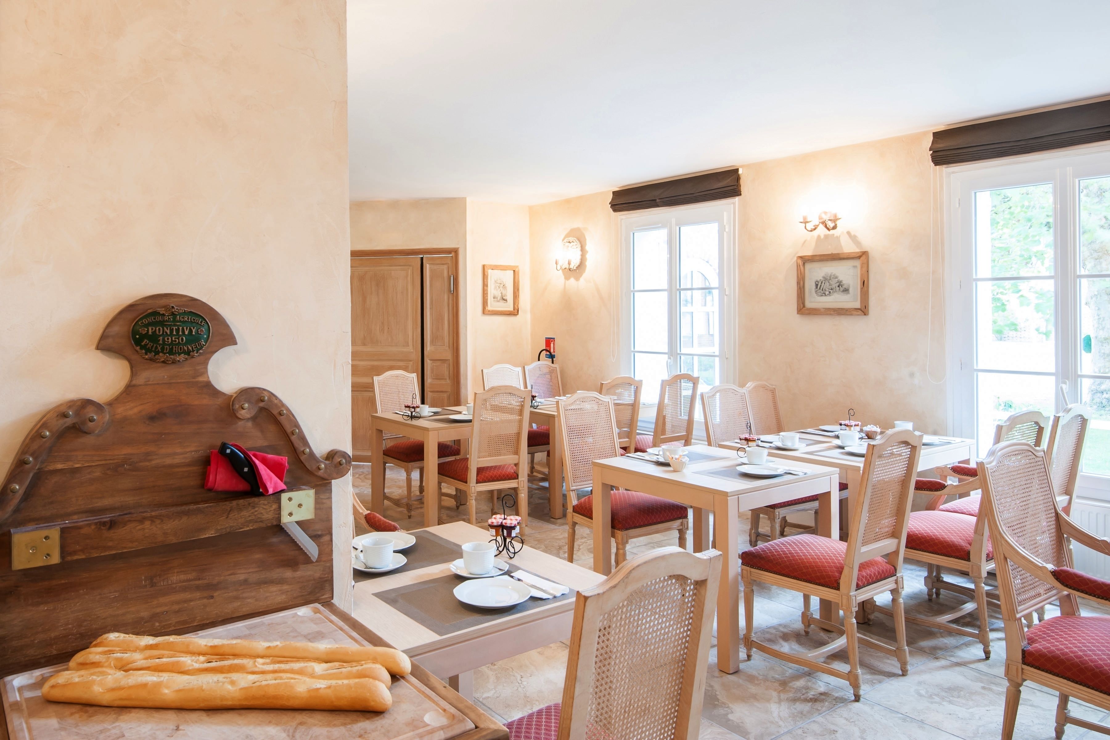 Un coin repas avec des tables et des chaises en bois, une tête de lit en bois ornée d’une plaque, et une fenêtre donnant sur un paysage verdoyant.