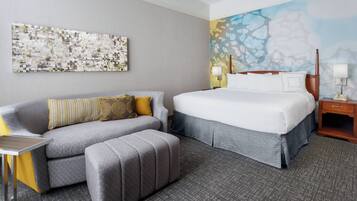 A hotel room with a grey sofa, a matching ottoman, a bed with white bedding, a nightstand with a lamp, and a wall with a textured abstract painting.