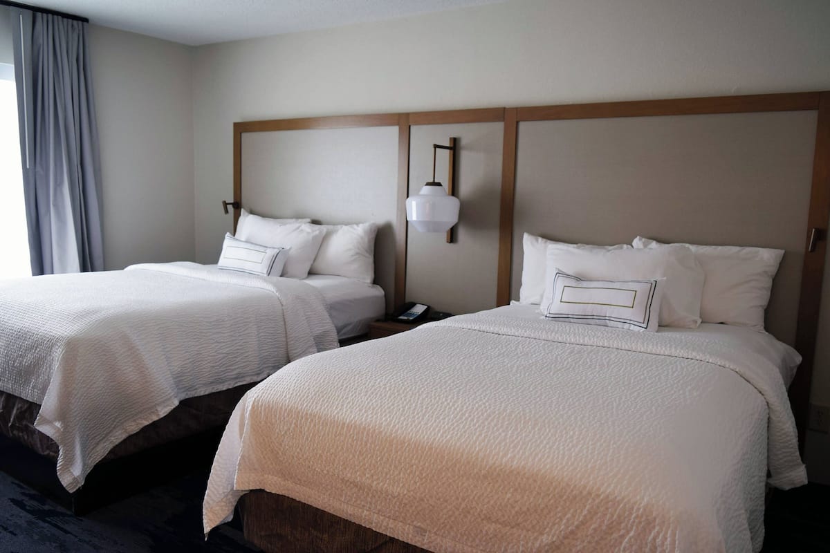 Two double beds with white bedding and a wooden headboard, a bedside table with a lamp, and a window with curtains.